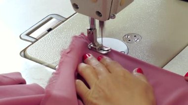 Kırmızı manikürlü, endüstriyel dikiş makinesinin iğnesinin altında pembe kumaşa rehberlik eden bir kadın elinin yakın çekimi.