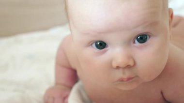Karnının üstüne uzanmış, geniş gözlü ve şaşkınlık dolu bir bebek resmi. Yumuşak ışıklandırma, sıcak tonlar ve doğal bir atmosfer çocukluğun duygularını ve saflığını yakalar. 4K