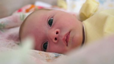 Yumuşak sarı elbiseli yeni doğmuş bir bebek, rahat bir battaniyenin üzerinde uzanmış, geniş, meraklı gözleriyle dünyaya bakıyor. Nazik renkler ve doğal ışık huzur verici ve iç açıcı bir atmosfer yaratır.