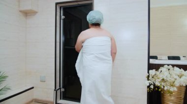 Havluya sarılmış ve şapka takmış son sınıf bir kadın saunaya doğru yürüyor. Sıcaklığı, sağlığı ve rahatlığı temsil eder..