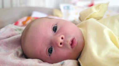 Yumuşak sarı bir elbiseye sarılı küçük bir bebek, desenli bir battaniyenin üzerinde uzanır, geniş bir alana bakar, merakla bakar. Yumuşak ışıklar ve ılık melodiler huzur verir