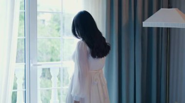 Zarif beyaz elbiseli bir kadın pencerenin yanında durur ve balkonu açar. Parlak, havadar atmosfer, hassas perdeler, doğal gün ışığı. Barış, sabah, uyum. 4K 'da çekilmiş..