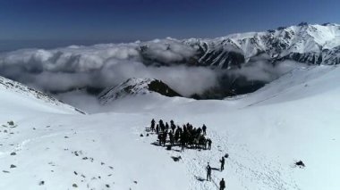 İnsansız hava aracı karlı bir dağ geçidinde duran bir grup yürüyüşçüden yükselip uzaklaşırken hava görüntüsü. Düşük tepelerde bulutlar süzülür dramatik alp manzarasıyla.