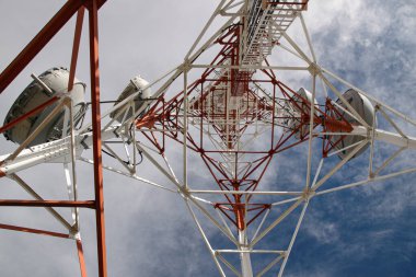 Eski antenli kırmızı ve beyaz iletişim kulesinin dinamik perspektifi, uydu antenleri, temeli beyaz bulutlarla kaplı, derin dağ mavisi gökyüzüne karşı. 