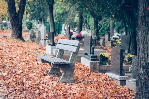 empty bench in the autumnal cemetery with colorful decorations on tombs and orange leaves on ground