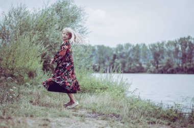 Güzel, tapılası ilahi kız, renkli uzun elbiseli, yeşil çayır elbiseli, nehrin kıyısında, vahşi kırsalda günün tadını çıkaran sihirli bir atmosfer.