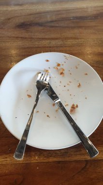 İçinde kırıntılar olan boş beyaz bir tabak ve ahşap bir masada kullanılmış çatal bıçak takımı. Bitmiş bir yemeği simgeliyor. Sahne lezzetli bir yemek deneyimi sonrasını yansıtıyor, memnuniyeti ve yemeğin sonunu vurguluyor.