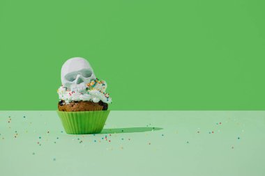 Krem şantili cadılar bayramı çöreği ve yeşil bir arka planda iskelet kafası. Ürkütücü bayram yemekleri konsepti. Canlı renkler arkaplanı.