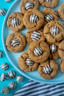 Close up of gingerbread blossom holiday cookies, stacked on blue round plate with gray striped napkins at bottom, with striped white chocolate pieces scattered around scene