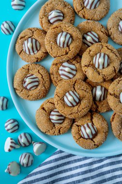 Close up of gingerbread blossom holiday cookies, with white chocolate striped candy on blue round plate with gray striped napkins at bottom, with striped white chocolate pieces scattered around scene