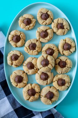 Peanut butter blossom cookies stacked on blue oval plate with black and white buffalo checked napkin on left side, on bright blue background