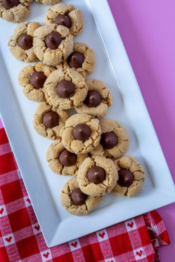 Plate of peanut butter blossom cookies, stacked on white rectangle plate with heart red and white checked napkin sitting on bright pink background