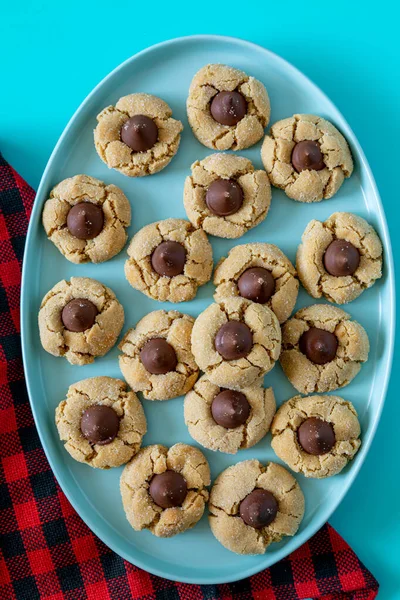 Peanut butter blossom cookies stacked on blue oval plate with red and black buffalo checked napkin on left side, on bright blue background
