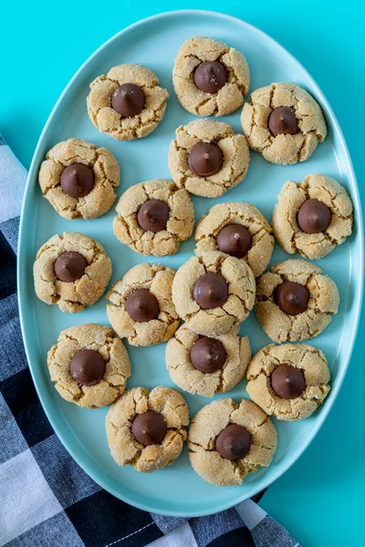 Peanut butter blossom cookies stacked on blue oval plate with black and white buffalo checked napkin on left side, on bright blue background