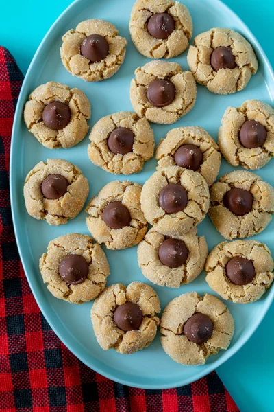 Close up of peanut butter blossom cookies stacked on blue oval plate with red and black buffalo checked napkin on left side, on bright blue background