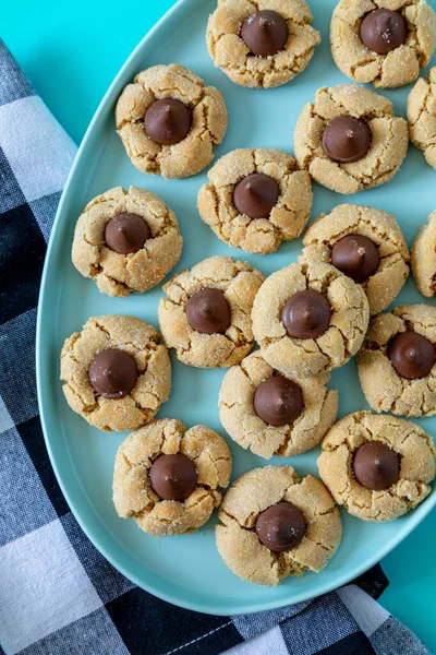 Close up of peanut butter blossom cookies stacked on blue oval plate with black and white buffalo checked napkin on left side, on bright blue background