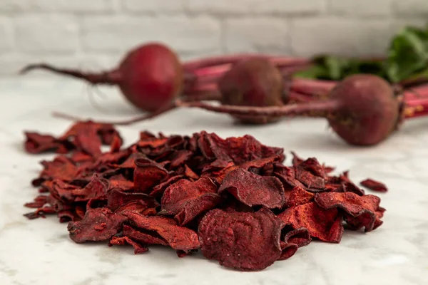 Red beet plant based chips sitting in a pile on marble kitchen counter with fresh beet roots in the background