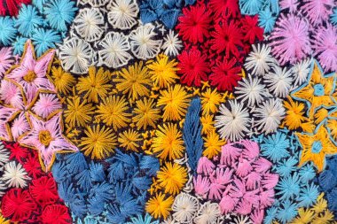 Bu resim renkli bir nakış deseninin yakın çekimini gösteriyor. Tasarım, kırmızı, sarı, mavi, pembe ve beyaz tonlarında çiçeklerle farklı çiçek motiflerinden oluşur. Çiçekler farklı dikiş teknikleri kullanılarak yapılır..