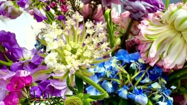 Bir buket çiçeğin parlak zoom videosu. Ana odak noktası karmaşık dalları olan soluk yeşil ve beyaz çiçekler. Çiçeklerin etrafında mor ve pembe tonlarda çeşitli çiçekler var. 