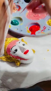 Bir kişi geleneksel dekoratif bir maske boyuyor. Maskenin beyaz bir yüzü, parlak siyah gözleri, parlak kırmızı dudakları ve karmaşık bir şekilde süslenmiş altın bir başlığı vardır. Sanatçı.