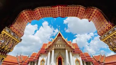 Turistler, Wat Benchamabophit 'in kemer yolundaki parlak mermer zeminde oturuyorlar, süslenmiş Tayland tapınağı binasına bakıyorlar, sırtları kameraya dönük, süslü dekore edilmiş altın rengi kapıdan geçiyorlar.