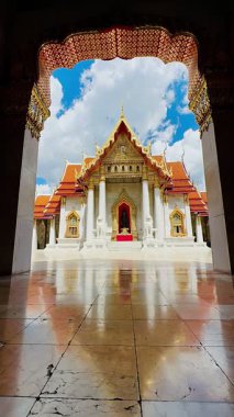 Turistler, Wat Benchamabophit 'in kemer yolundaki parlak mermer zeminde oturuyorlar, süslenmiş Tayland tapınağı binasına bakıyorlar, sırtları kameraya dönük, süslü dekore edilmiş altın rengi kapıdan geçiyorlar.