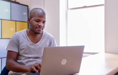 Çekici genç siyahi erkek ofis yöneticisi gündelik tişörtle dizüstü bilgisayarda blog okuyor, geniş pencereli modern çalışma alanında meslektaşlarını bekliyor.