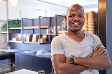 Yakışıklı, arkadaş canlısı, gülümseyen, sıradan giyinmiş modern ofis dinlenme odasının kafesinde duran, kollarını kavuşturup kameraya neşeyle gülümseyen siyahi bir adamın portresi.