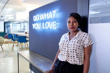 Empowering portrait of proud black professional curvy woman standing with arms crossed wearing pattern blouse, in front of trendy neon sign in modern office room clipart