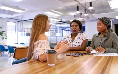 Candid shot of diverse only women group of colleagues working at large wooden table with coffee cup and bright lights, in modern office break room clipart