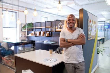 Yakışıklı, arkadaş canlısı, gülümseyen, sıradan giyinmiş modern ofis dinlenme odasının kafesinde duran, kollarını kavuşturup kameraya neşeyle gülümseyen siyahi bir adamın portresi.