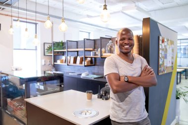 Yakışıklı, arkadaş canlısı, gülümseyen, sıradan giyinmiş modern ofis dinlenme odasının kafesinde duran, kollarını kavuşturup kameraya neşeyle gülümseyen siyahi bir adamın portresi.