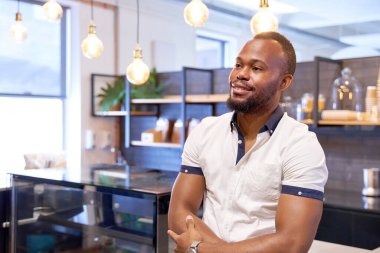 Yakışıklı, arkadaş canlısı, gülümseyen, sıradan giyinmiş modern ofis dinlenme odasının kafesinde duran, kollarını kavuşturup kameraya neşeyle gülümseyen siyahi bir adamın portresi.