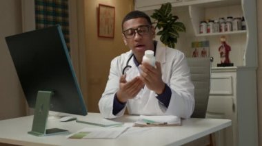 Doktor hastayla çevrimiçi konuşuyor. Afro-Amerikalı doktor hastane ofisinde bilgisayar monitörüne ilaç şişesini gösteriyor. Tıbbi danışmanlık çevrimiçi. 4K