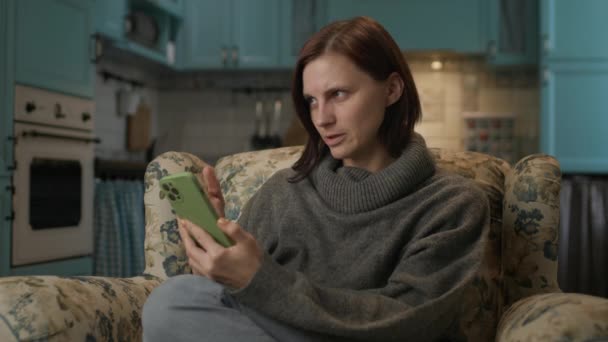 携帯電話でネットサーフィンする女 家に座って携帯電話で30代の笑顔の女性 — ストック動画