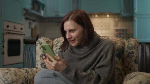 携帯電話を使用して驚きの女性 自宅の居心地の良いソファに座って携帯電話で良いニュースを読んで非常に興奮30代の女性 — ストック動画