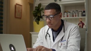 Doktor dizüstü bilgisayar kullanıyor. Afro-Amerikalı pratisyen hastane ofisinde çalışan dizüstü bilgisayarda daktilo kullanıyor. 4K