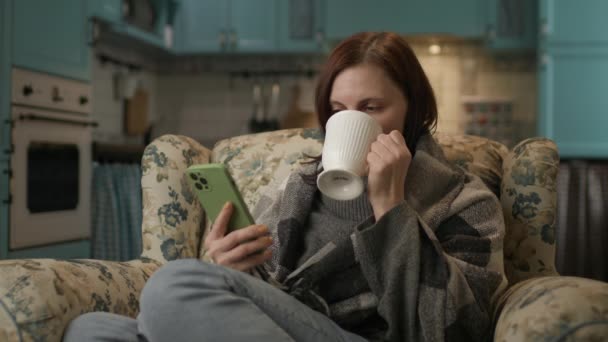 自宅で携帯電話を使ってネットサーフィンをしている女性 居心地の良いソファに座って熱いお茶を飲むスマートフォンを使って30代の笑顔の女性 — ストック動画