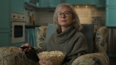 Kıdemli Kadın TV izliyor ve patlamış mısır yiyor. Olgun bir bayan, evdeki koltukta oturmuş komik şovlar izlerken gülümsüyor. 4K