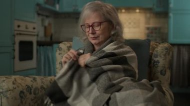 Yaşlı kadın evde üşüyormuş. Koltukta oturan, battaniyeye sarılmış olgun bir kadın. 4K