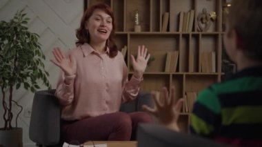 Çocuk terapisti, psikolog ofisinde oturan bir çocukla şarkı söyleyip oyunlar oynuyor. Çocukların zihinsel sağlıklarının farkındalığı. Omuz manzarasının üstünden. 4K