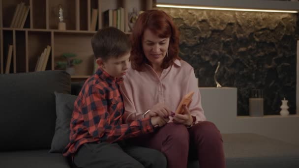 母は自閉症の息子に自宅のソファに座ってスマートフォンを使用する方法を学びます 母親の助けを借りて携帯電話を使って自閉症の少年 — ストック動画