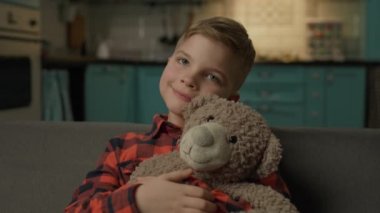 Tatlı çocuk evde oyuncak ayıya sarılıyor. Öğrenci çocuk, sevimli ayı oyuncağıyla kanepede oturmuş kameraya bakıyor. 4K