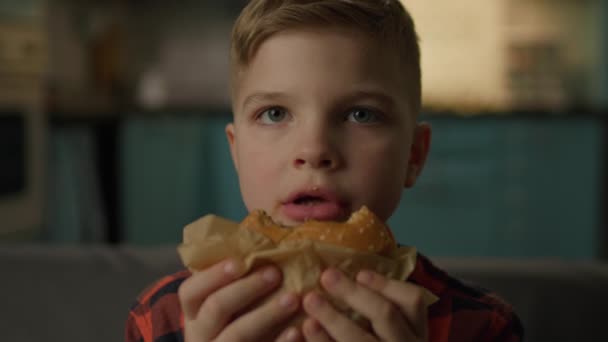 饥饿的孩子吃汉堡看着相机 学童坐在家里的沙发上咀嚼快餐汉堡 关门了 — 图库视频影像