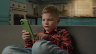 Evde cep telefonu kullanmaktan sıkılmış çocuk. Kanepede tek başına oturan akıllı telefonu kullanarak internette sörf yapan hoşnutsuz çocuk. 4K