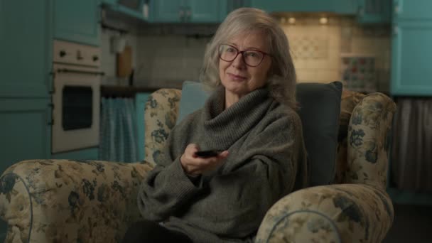 60年代的女人看电视 老太太抱着遥控器 坐在家里的扶手椅上看电视节目 — 图库视频影像