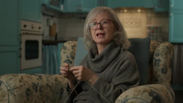成熟女子编织在家看电视 老太太喜欢坐在舒适的扶手椅上织毛衣 — 图库视频影像
