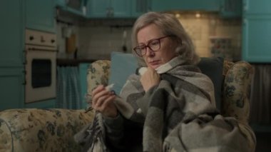 Hasta Olgun Kadının Ateşi Var. Evdeki koltukta oturan, termometreyi elinde tutan hasta yaşlı bir kadın. 4K
