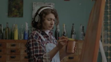 Cheerful Artist Creating Her Masterpiece Listening To Music. Female painter in headphones painting on canvas in workshop. Happy woman working in art space. High quality 4k footage