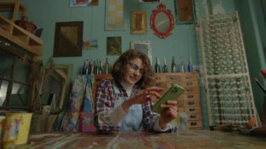 Atölyede oturan akıllı telefonu kullanan kadın mesajlaşması. Genç yaratıcı sanatçı cep telefonuyla internette sörf yapıyor. 4K